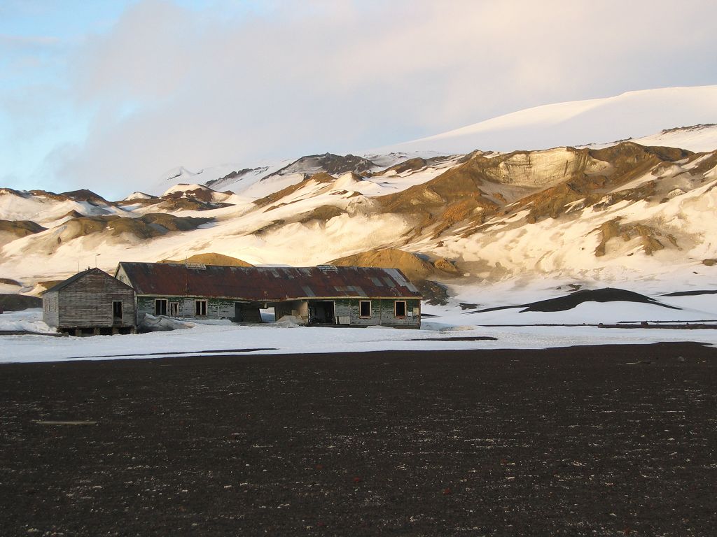 An old building on Deception Island is in shade as the long sunset lights the slopes above.  Click for next photo.